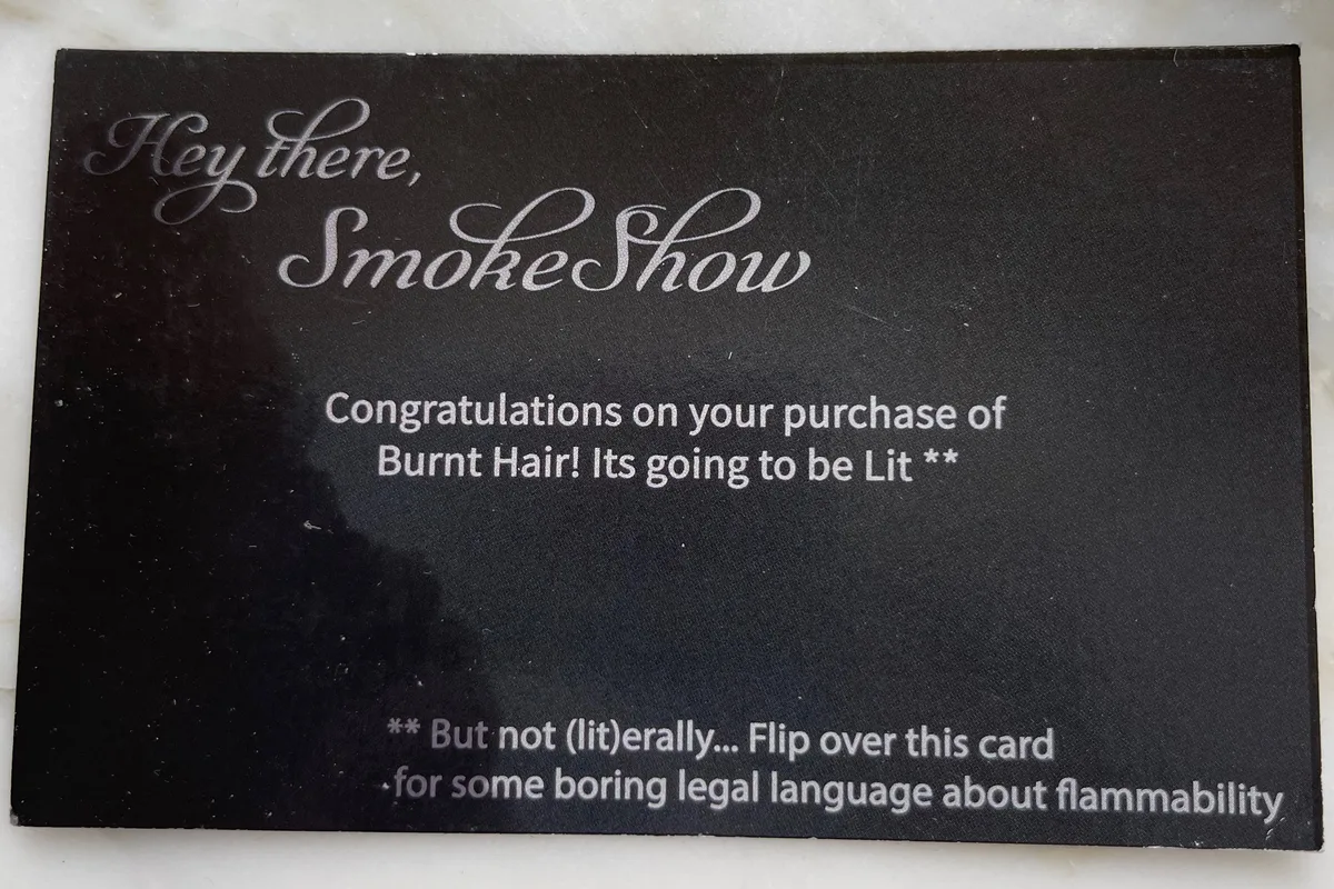 A small black card from inside the Burnt Hair perfume box, printed with cursive silver lettering that reads 'Hey there, SmokeShow — Congratulations on your purchase of Burnt Hair! Its going to be Lit ** ** But not (lit)erally… Flip over this card for some boring legal language about flammability.'